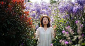 背の高い紫色の雑草(マツバウンラン、クサフジ、ノアザミなど)の中に立つ日本人の女性