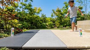庭の明るい日差しの中、濃いグレー、薄いグレー、テラコッタ風ベージュの3種類の屋外用タイルを並べた比較。表面の質感や色の違いがわかり、裸足の子供がベージュのタイルに足を乗せている。