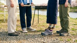 砂利道と草地が混在する河原のバーベキュー会場で、足元をしっかりと守る丈夫なアウトドアシューズを履いた女子大学生。安定感のある靴で転倒のリスクを回避している。