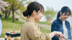 バーベキュー中に、崩れにくいお団子ヘアと編み込みスタイルにまとめた女子大学生の横顔。髪が料理に入らないよう清潔感を保ち、調理中も作業に集中している。