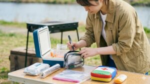 バーベキュー会場のピクニックテーブルで、除菌ウェットティッシュ、絆創膏、油性マジック、アルミホイル、ジップロックなどの便利な持ち物を整理する女子大学生。周囲への配慮と準備の良さを示している。